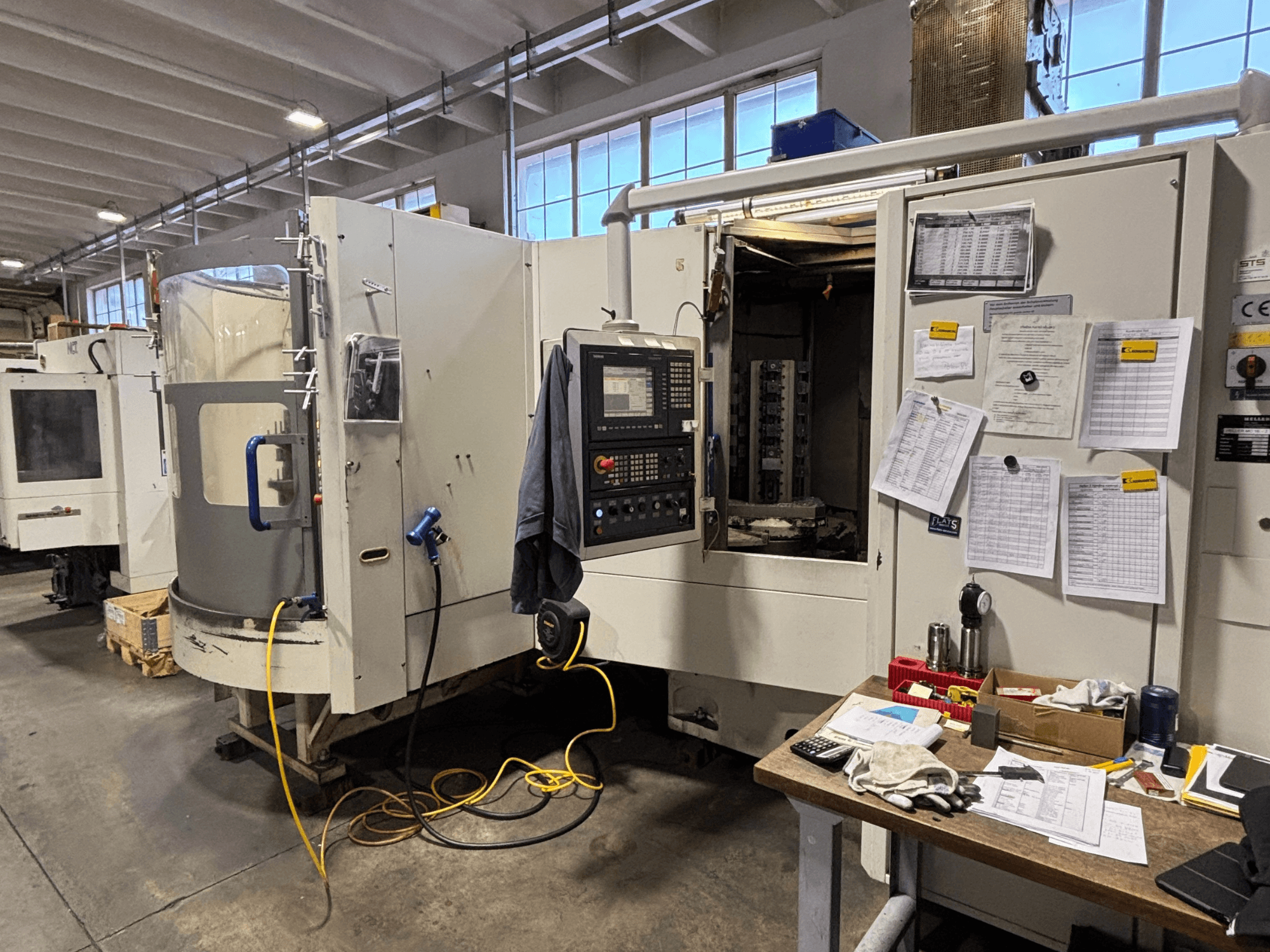 CNC machine in a workshop, front view, featuring control panel and documentation on the side; tools and materials on the table.