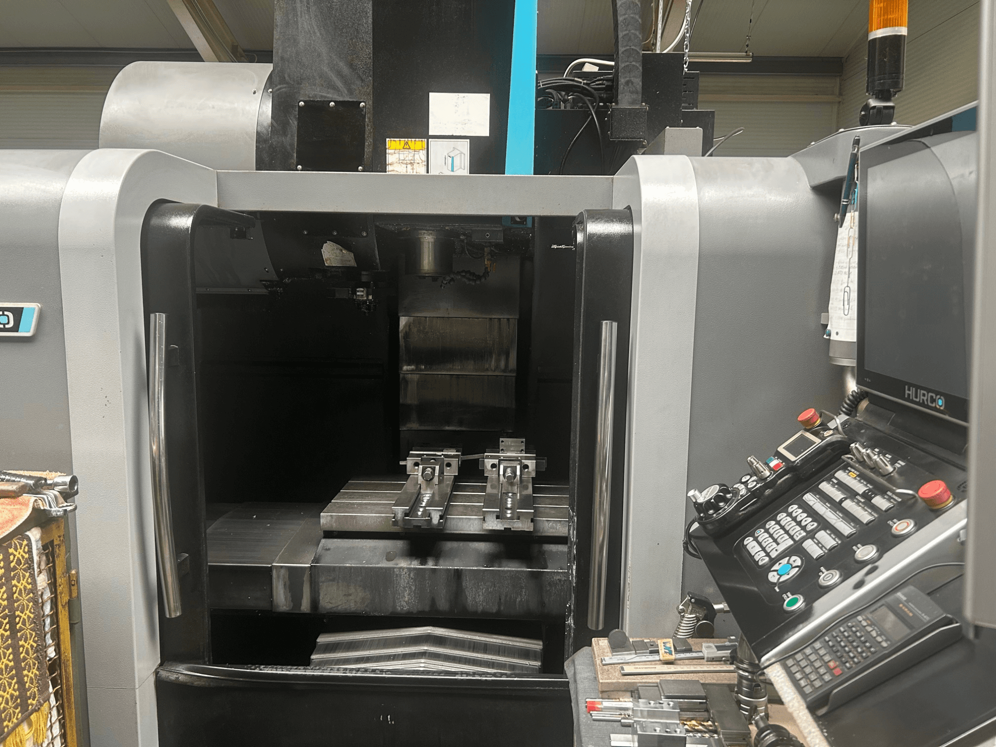Hurco CNC machining center front view, showing tool holder, metal workpiece, and control panel with buttons and display.