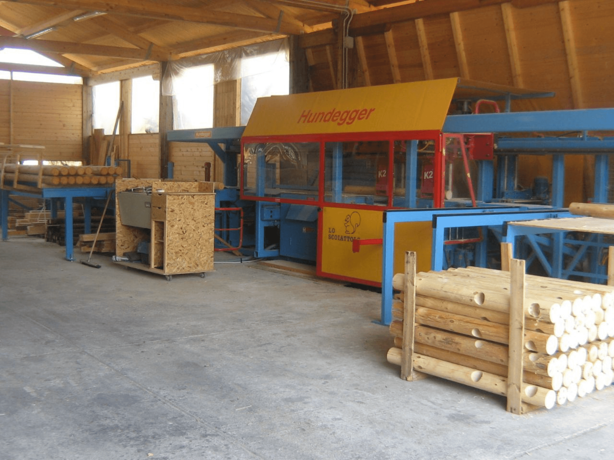 Hundegger woodworking machine in a workshop, side view, with wooden logs and processed timber stacked nearby.