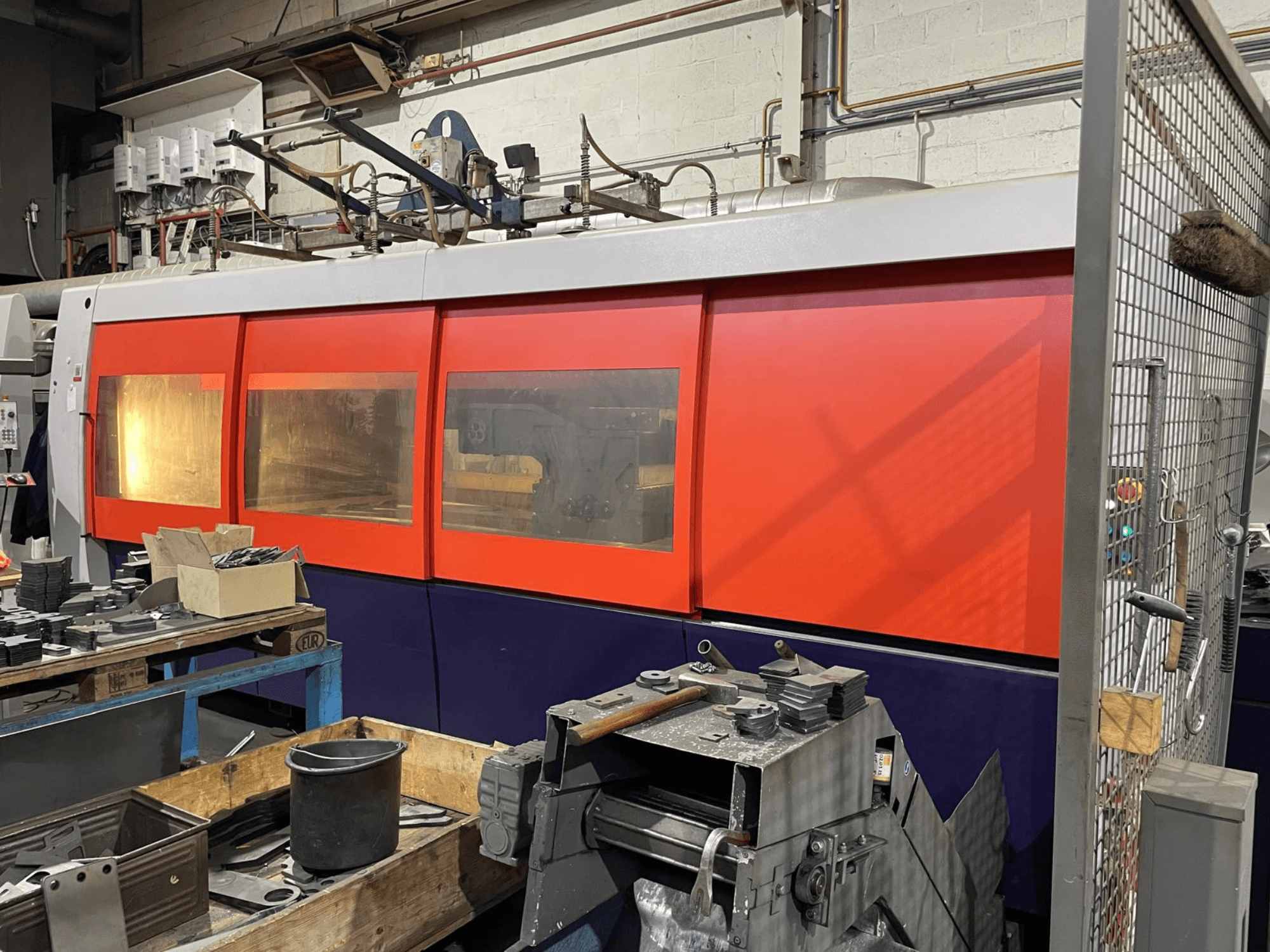 Close-up of a orange and white industrial laser cutting machine, side view, with control panel and workshop tools in foreground.