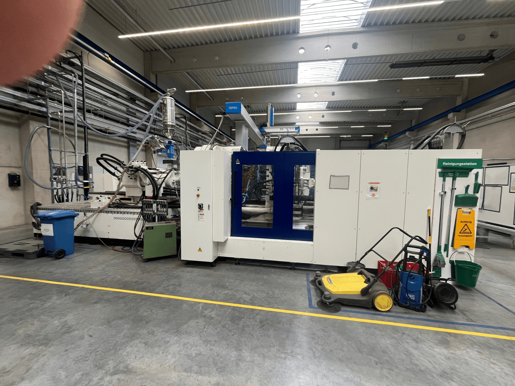 Industrial machine in a factory, front view, with blue and white panels, hoses, and cleaning equipment nearby.