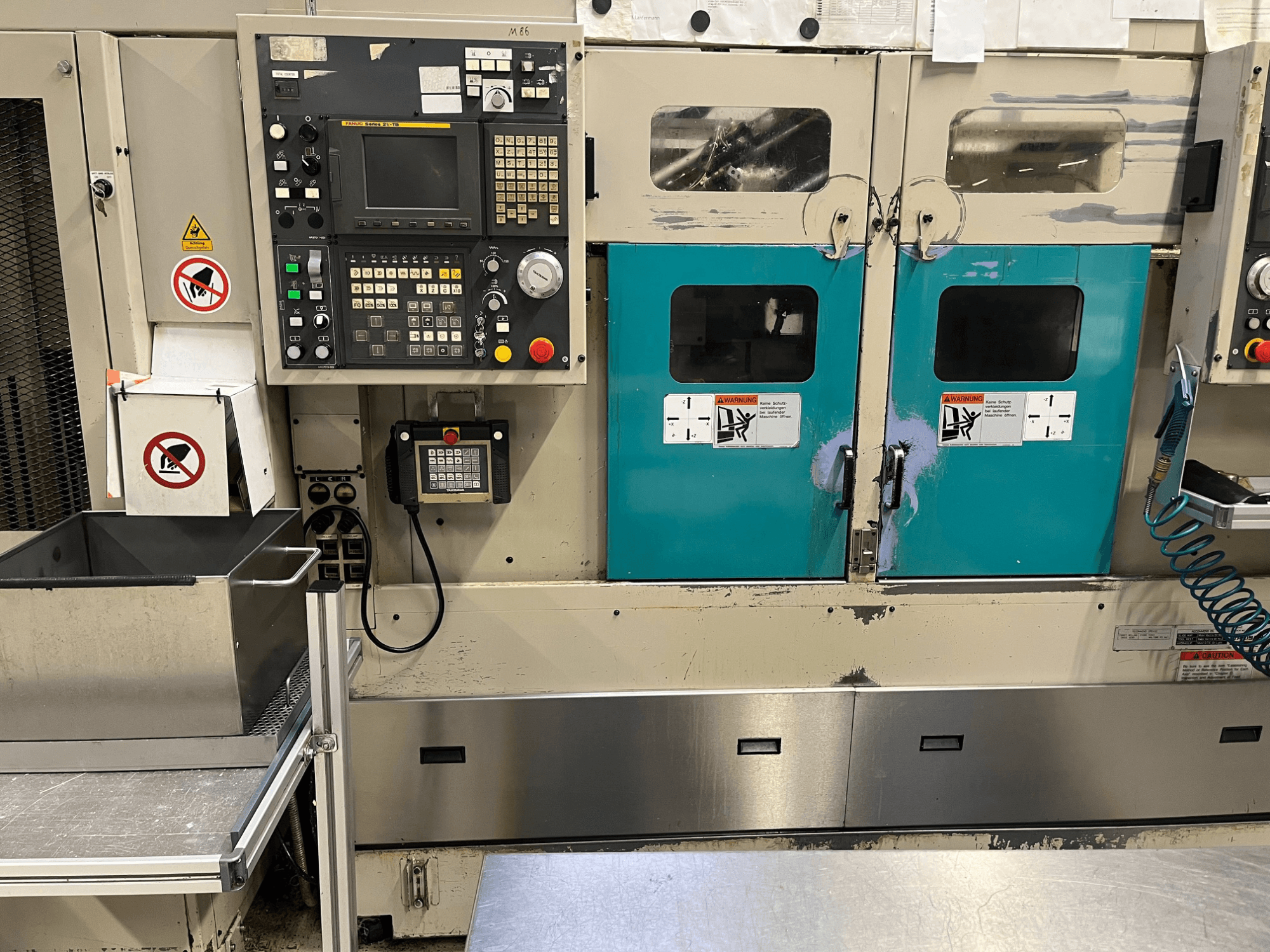 Control panel of a manufacturing machine with teal doors, safety warnings, and a keypad; front view, stainless steel finish.