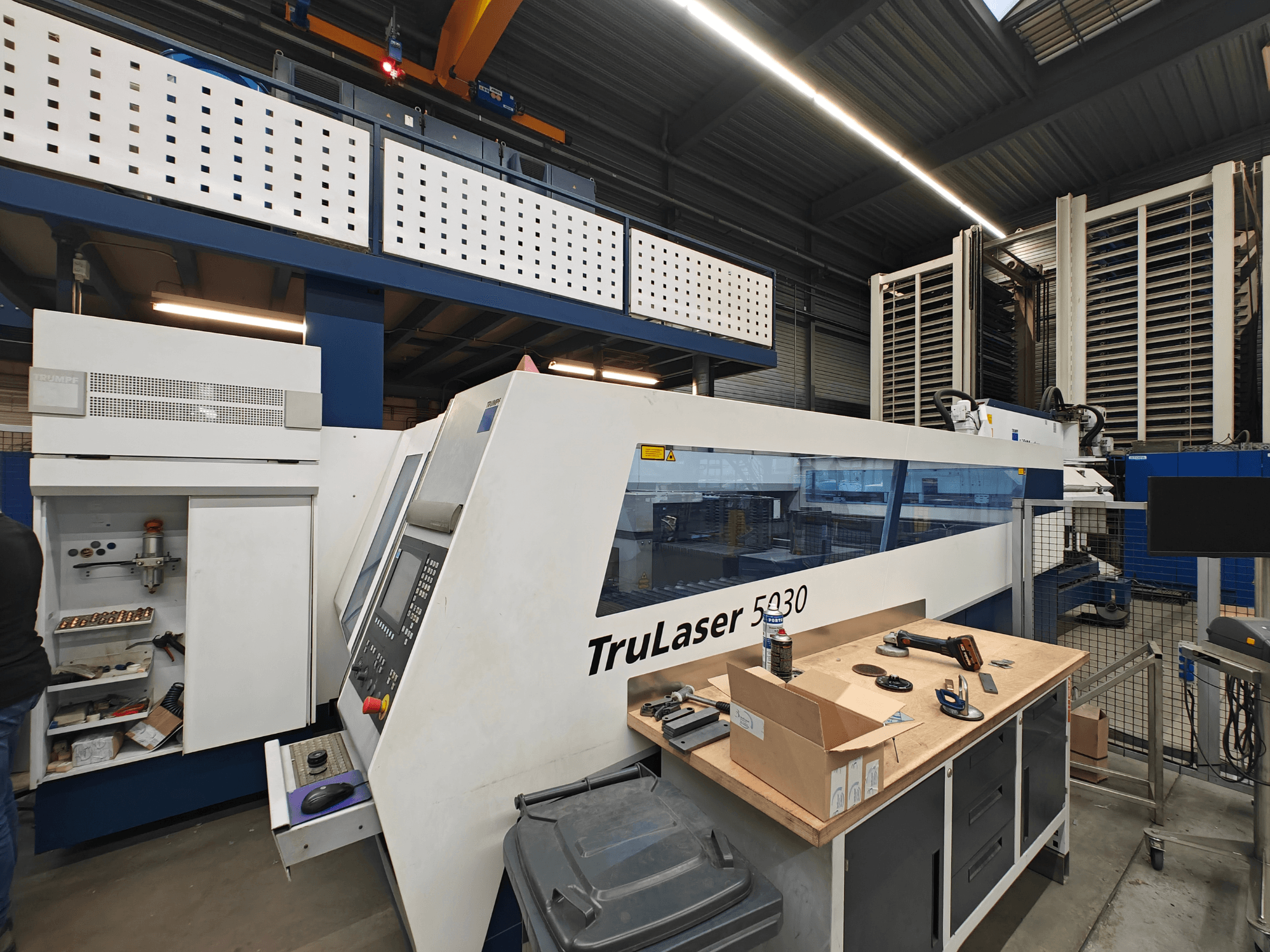 TruLaser 5030 machine in a workshop, front view with control panel visible, surrounded by tools and equipment.