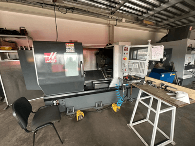 Haas CNC lathe (model ST-10) in a workshop, side view showing controls, tools on a workbench, and a chair nearby.