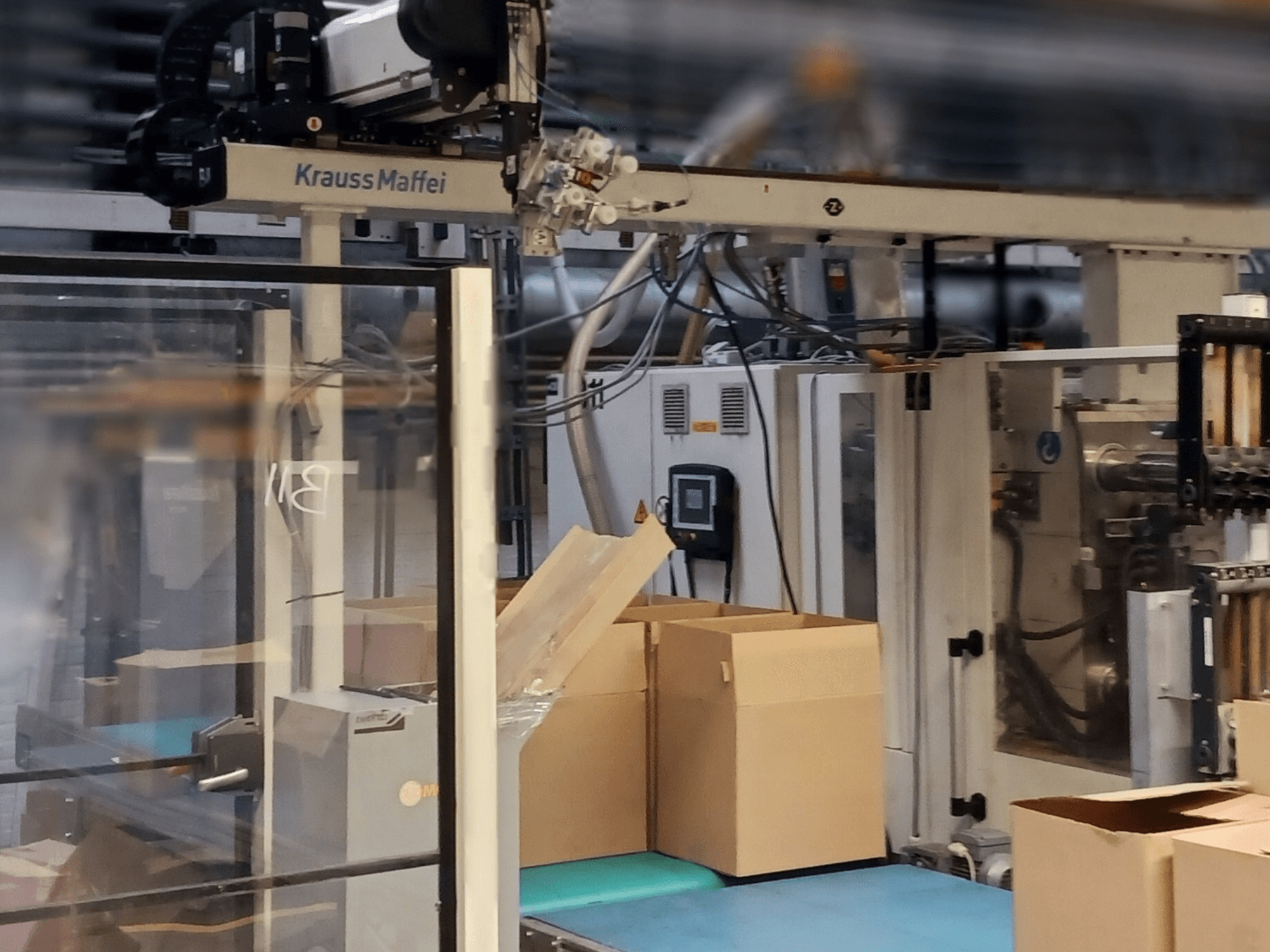 Krauss Maffei automation system in a factory, front view showing boxes on a conveyor and machinery components in operation.