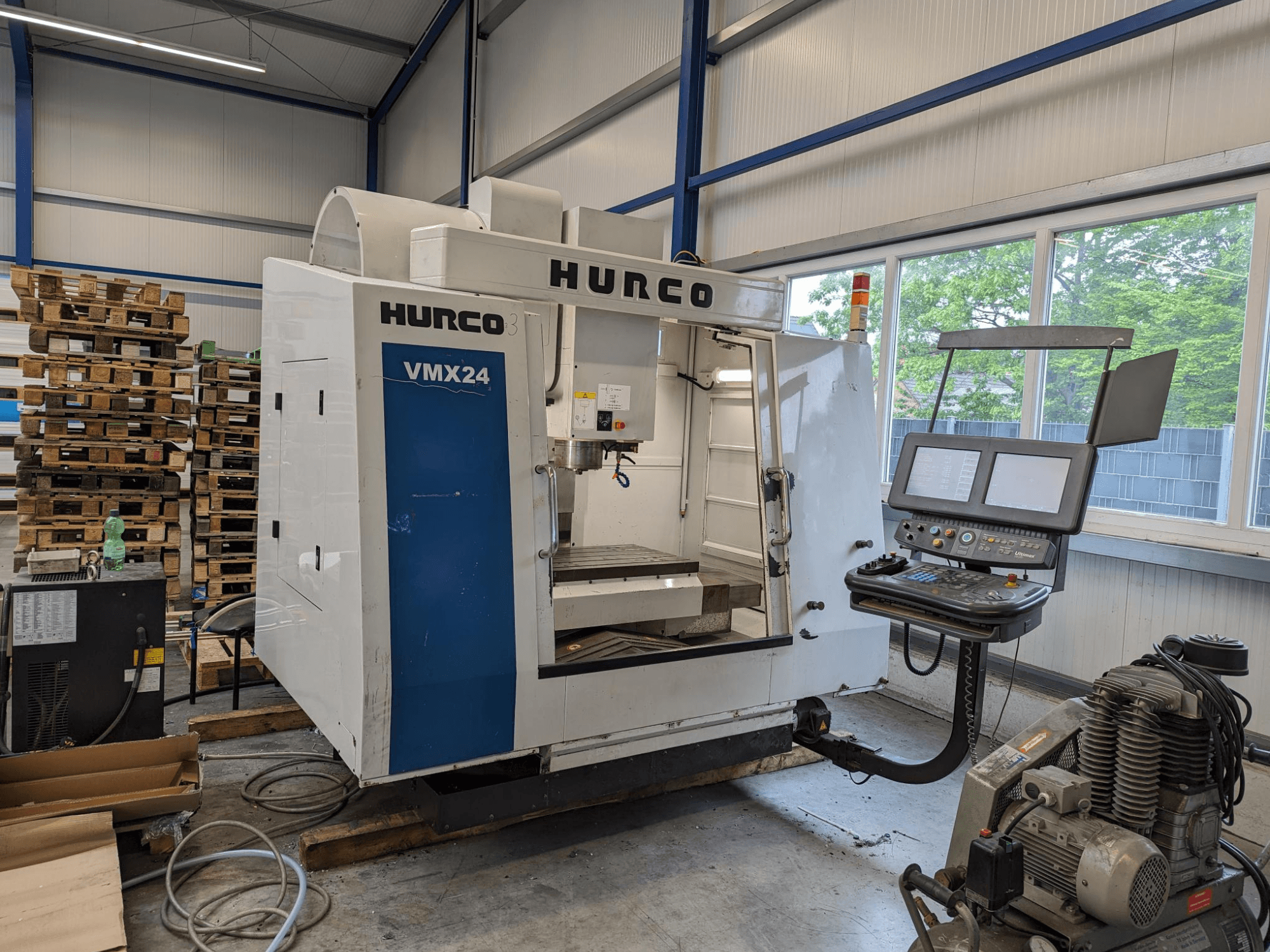 Hurco VMX24 milling machine, front view, with control panel and tool area; stacked pallets in background; industrial workspace setting.