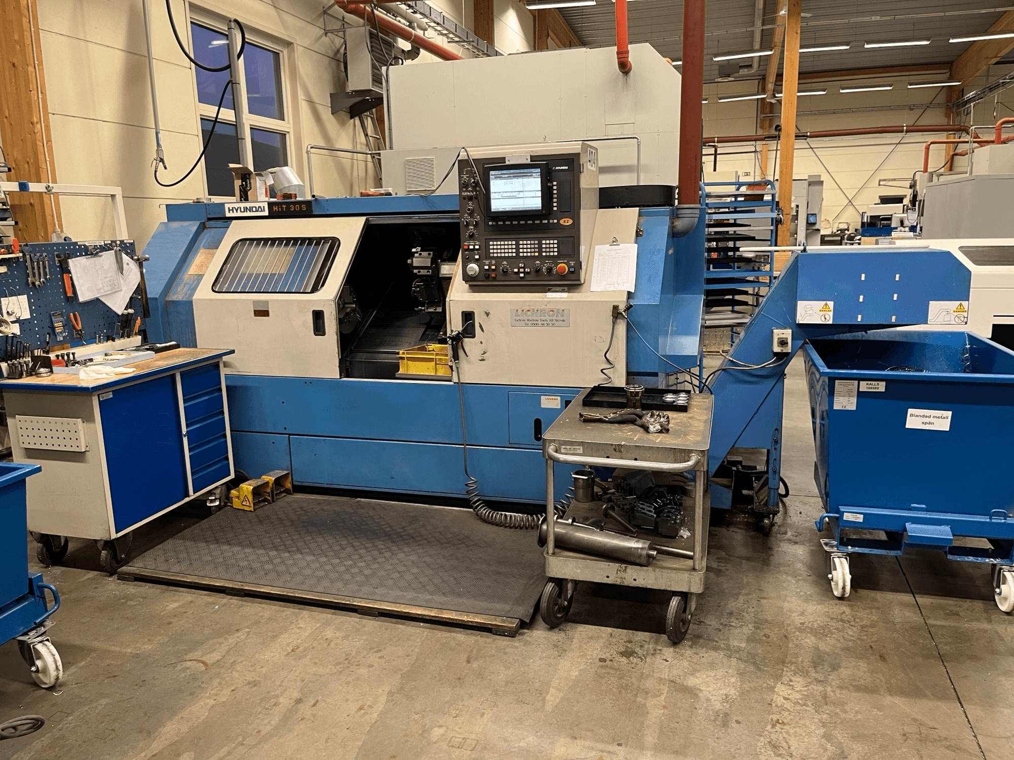 Hyundai HIT 30S horizontal turning machine, front view, with control panel and tools on a nearby cart in a workshop setting.