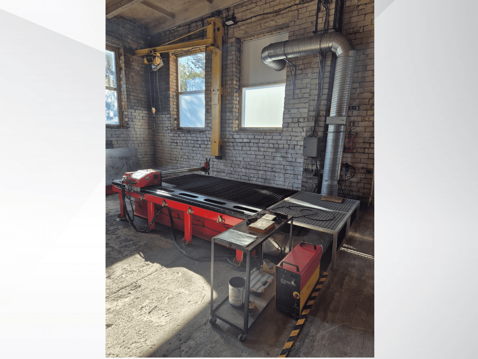 Red CNC plasma cutting table with control panel and metal exhaust duct, viewed from the side in a well-lit workshop.