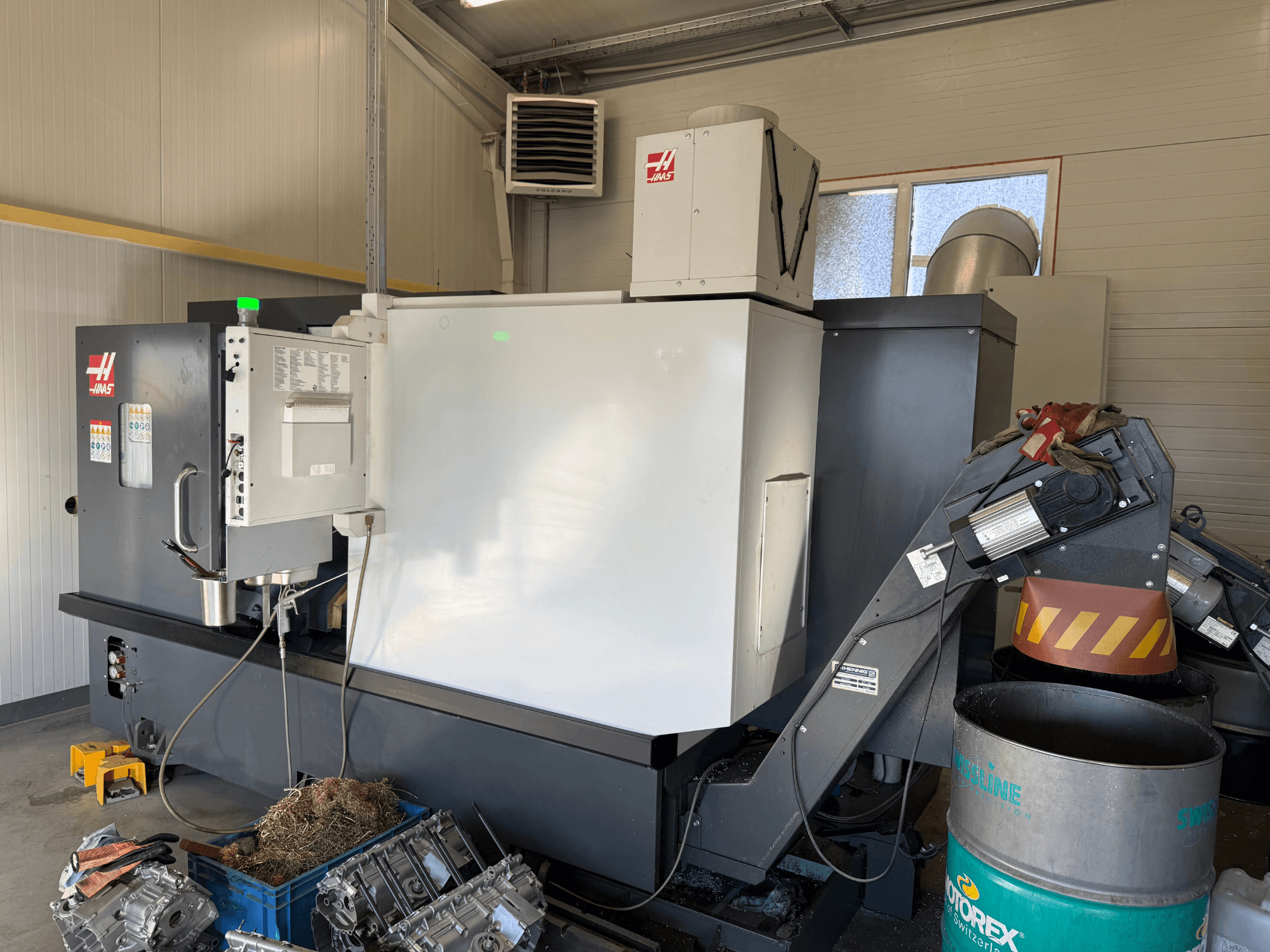 Haas CNC machine with control panel, side view; visible coolant and oil lines; metal scraps in blue bin; industrial setting.