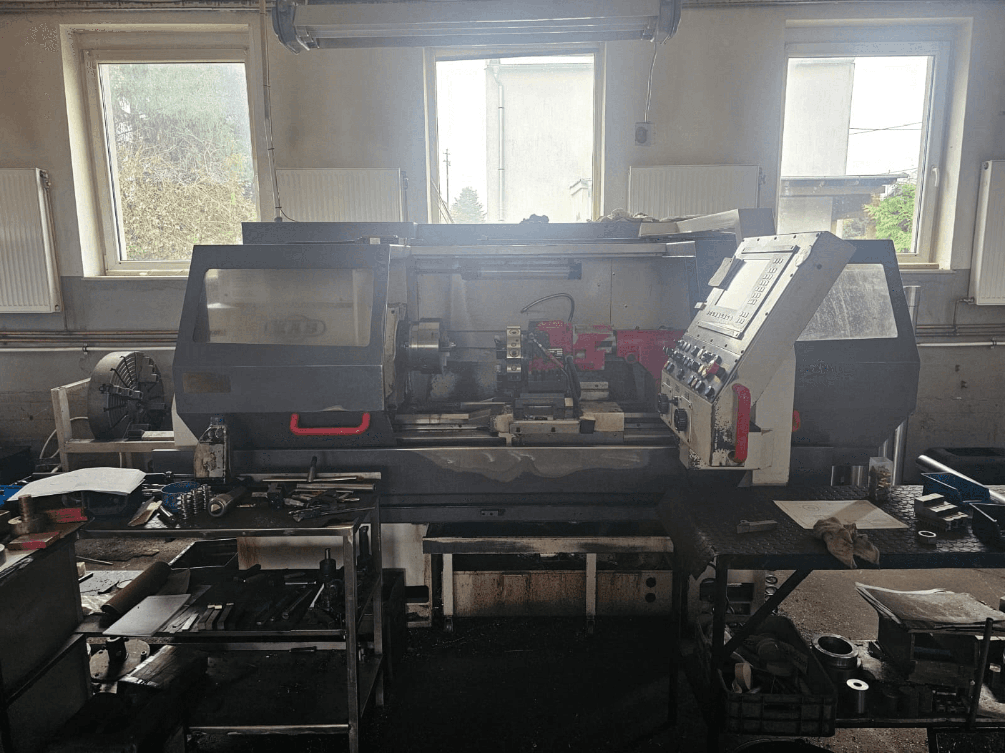 Lathe machine in a workshop, front view, with control panel and tools on tables, surrounded by windows and cluttered workspace.