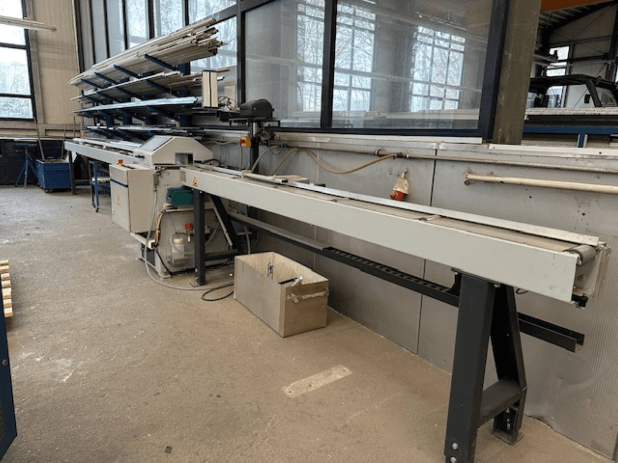 SCHÜCO PGS Circular Metal Saw in a workshop, side view with an adjustable cutting bed and a shelf of metal profiles above.