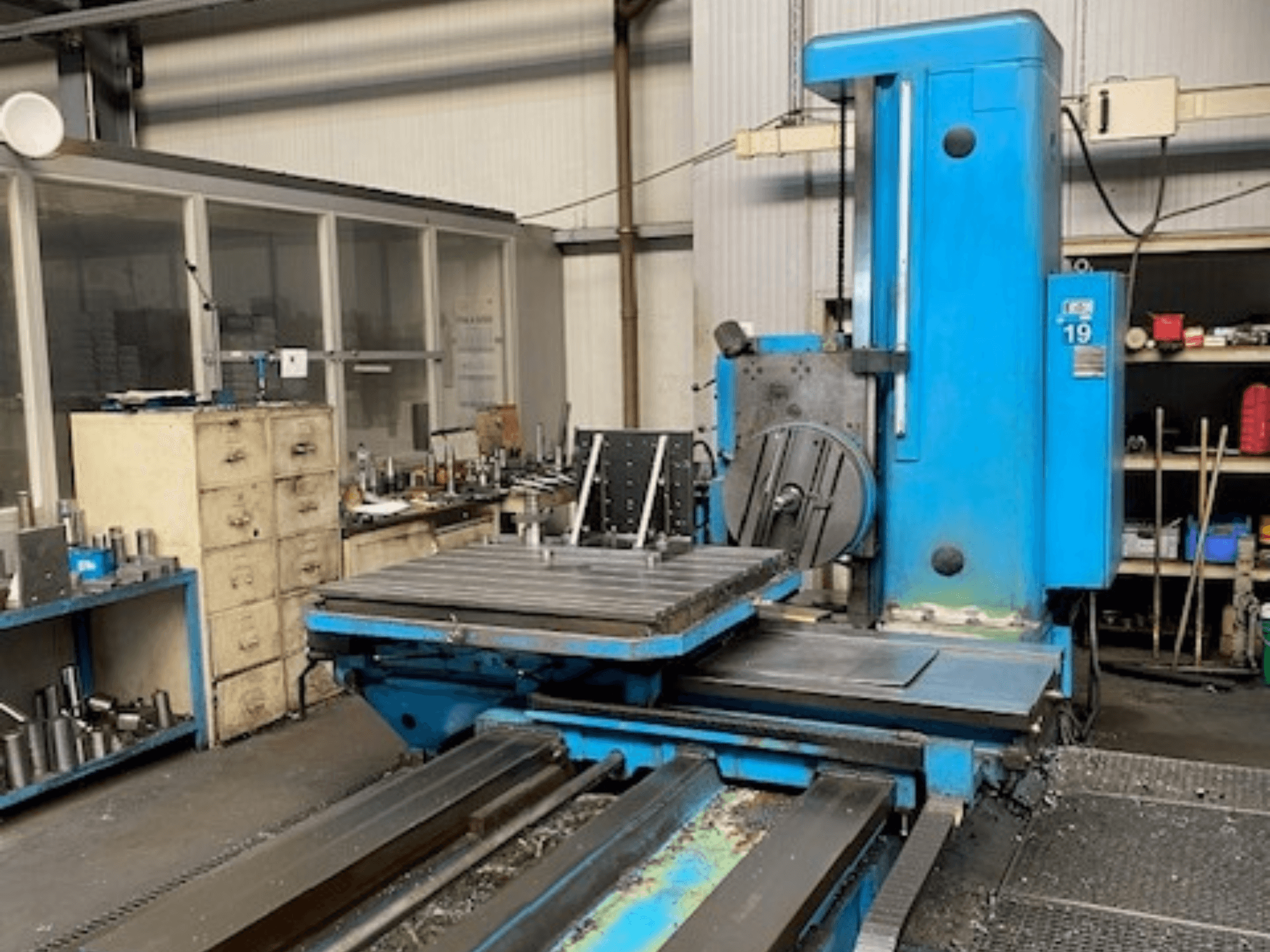 Blue milling machine in front view, model number 19, on a workshop floor with tools and a storage cabinet in the background.