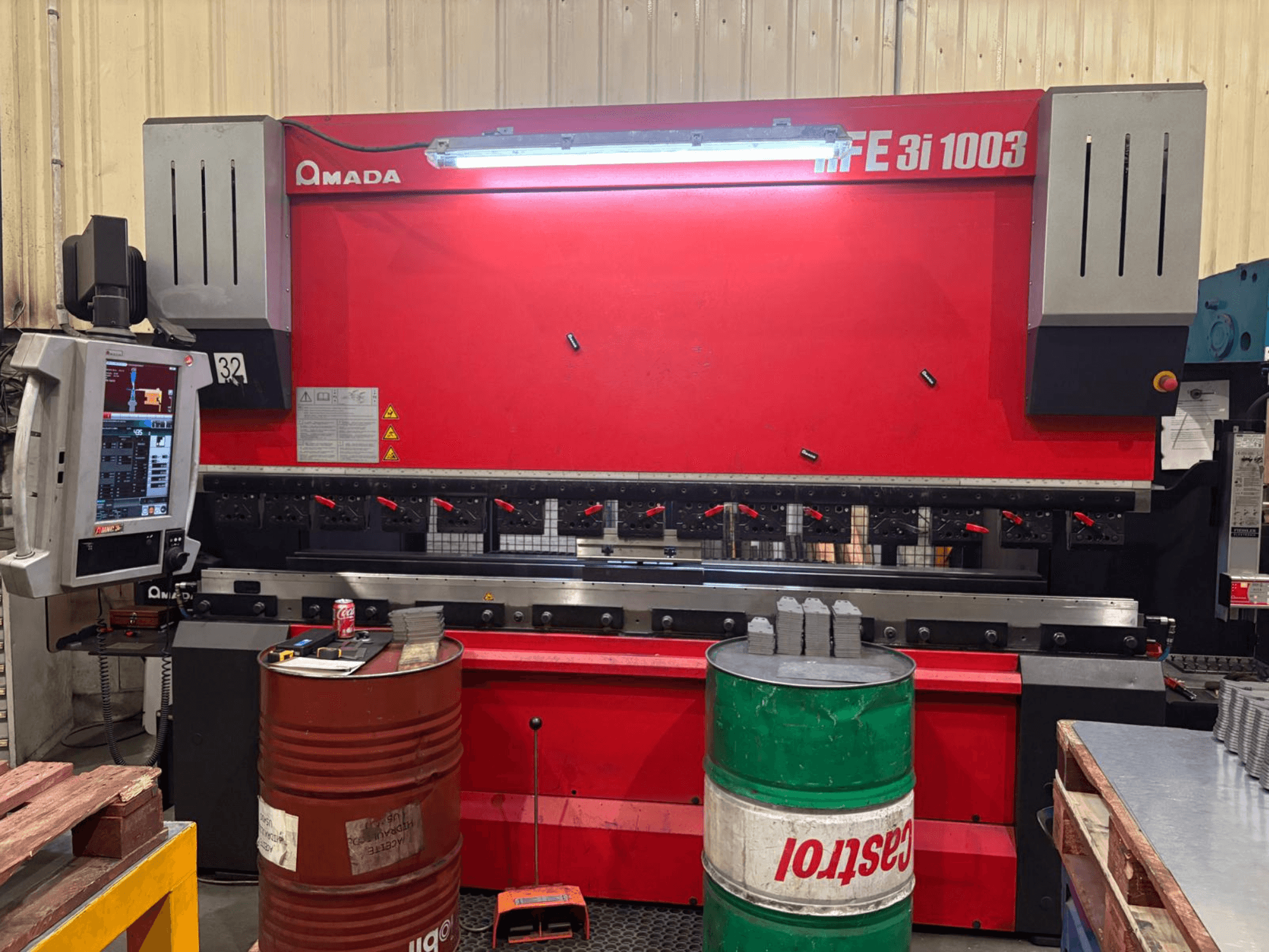 Amada HFE 3i 1003 press brake in a workshop, featuring a control panel, metal workpiece, and two barrels nearby.