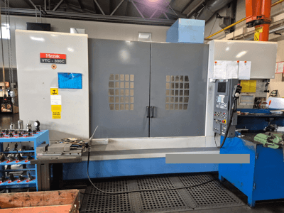 Mazak VTC-300C machine in a factory, front view showing control panel, grey doors with window grid, and nearby equipment.