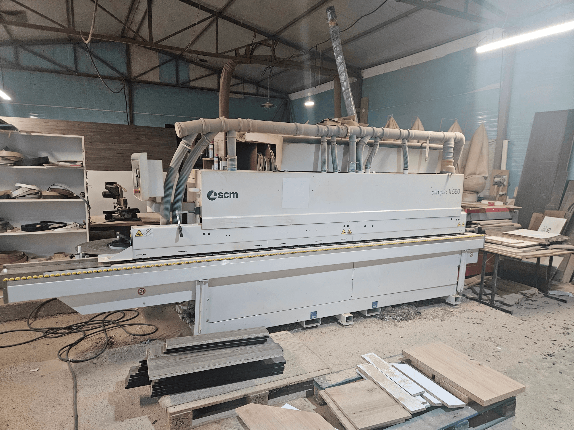 SCM Olimpic K560 edge banding machine in a workshop, front view, with wood panels and tools visible on the floor.