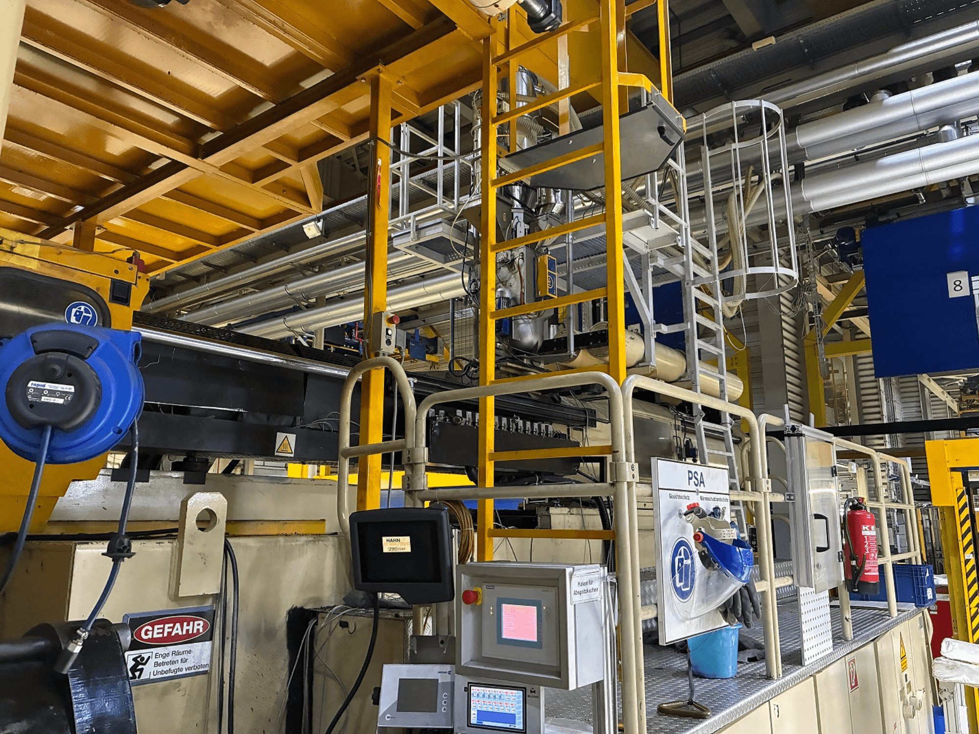 Industrial machinery control panel with blue safety equipment and yellow scaffolding, side view showing pipes and warning signs.