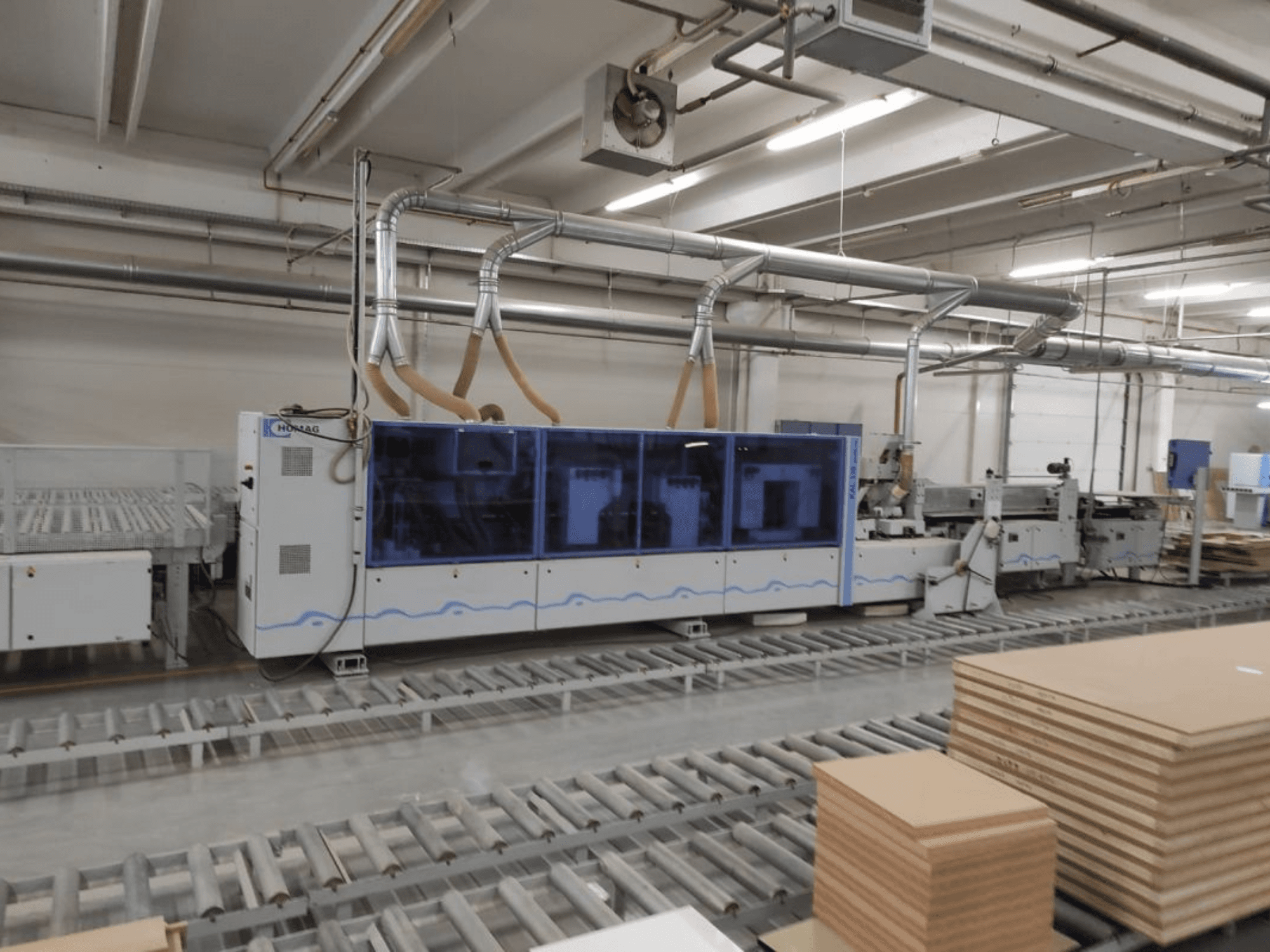 HOMAG production machine in a factory, front view, featuring blue casing and conveyor system with stacked plywood sheets.