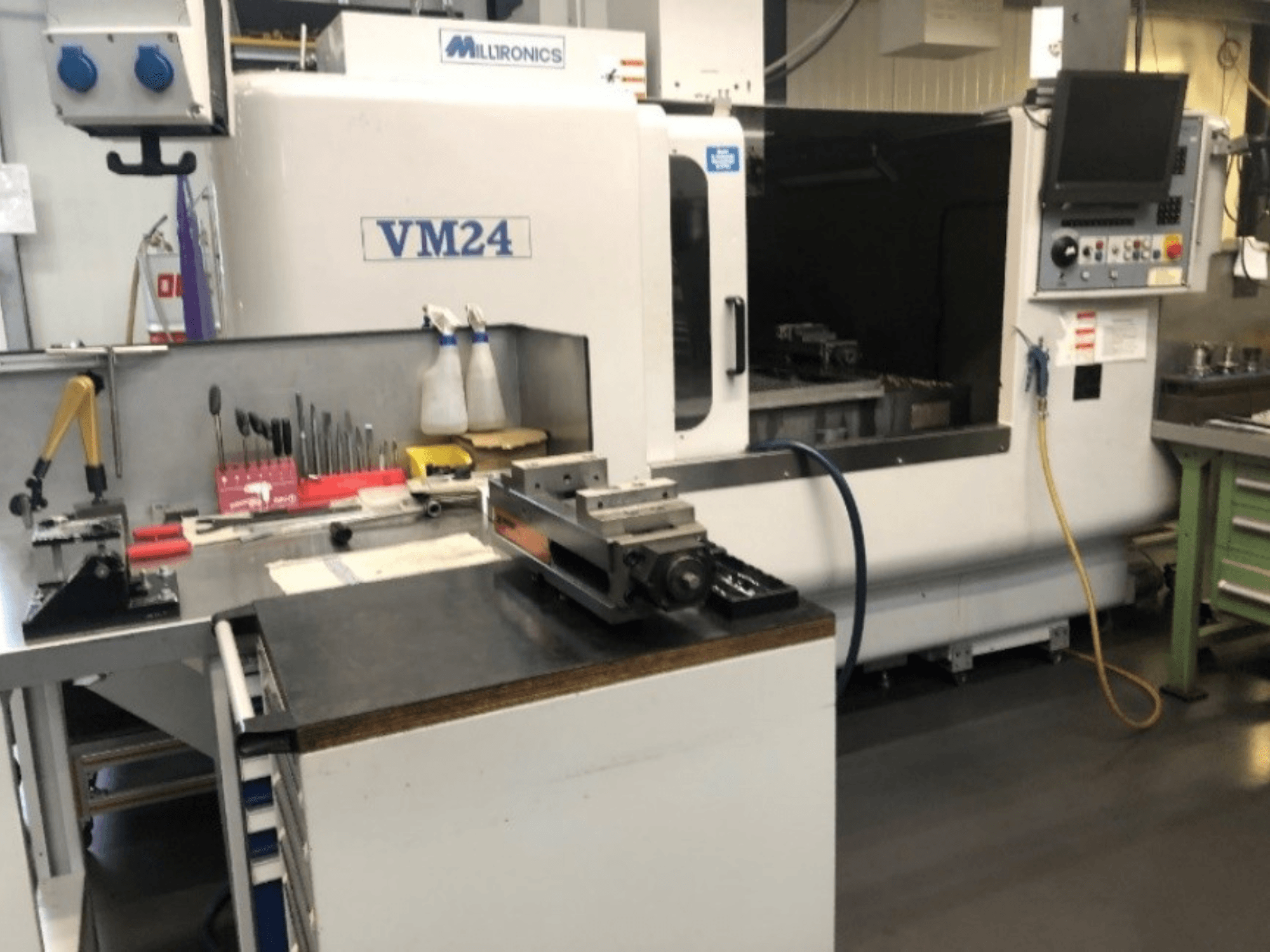 Machinery workspace featuring the Milltronics VM24 CNC machine. Front view with tools and equipment on a workbench.