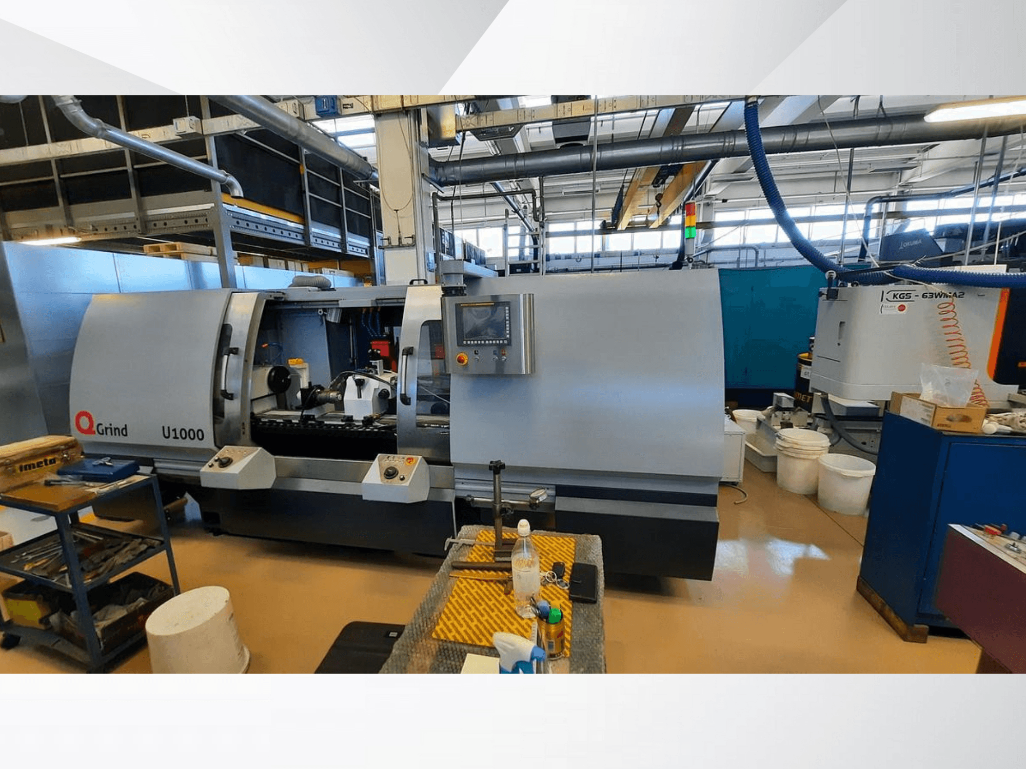 Q-Grind U1000 machine in a workshop, showcasing a front view with control panel and open cutting area, surrounded by equipment.