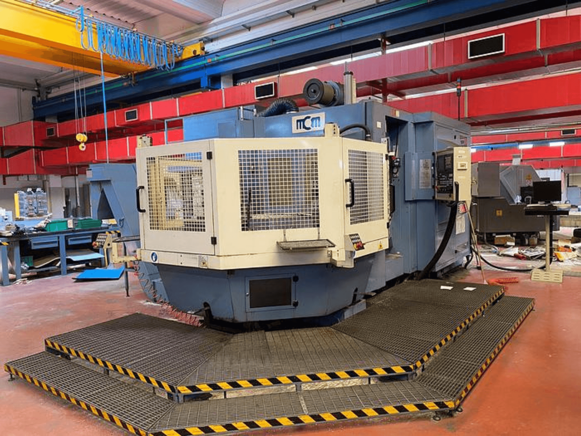 MCM CNC machine in a factory, front view with protective cage, black and yellow safety edges, surrounded by industrial equipment.
