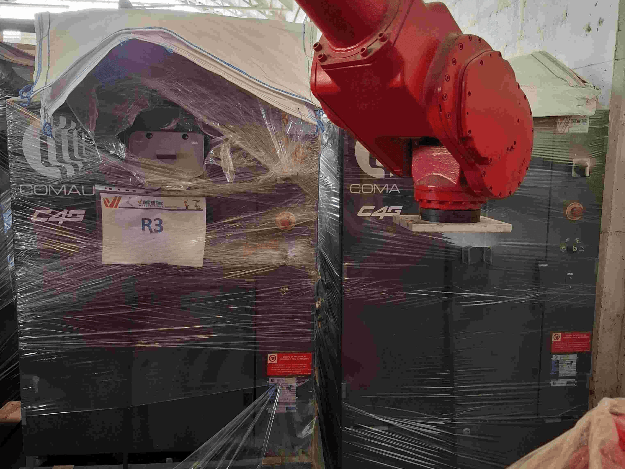 Comau C4G robotic arm seen from the front, wrapped in plastic, labeled "R3," with caution stickers visible on the machine.