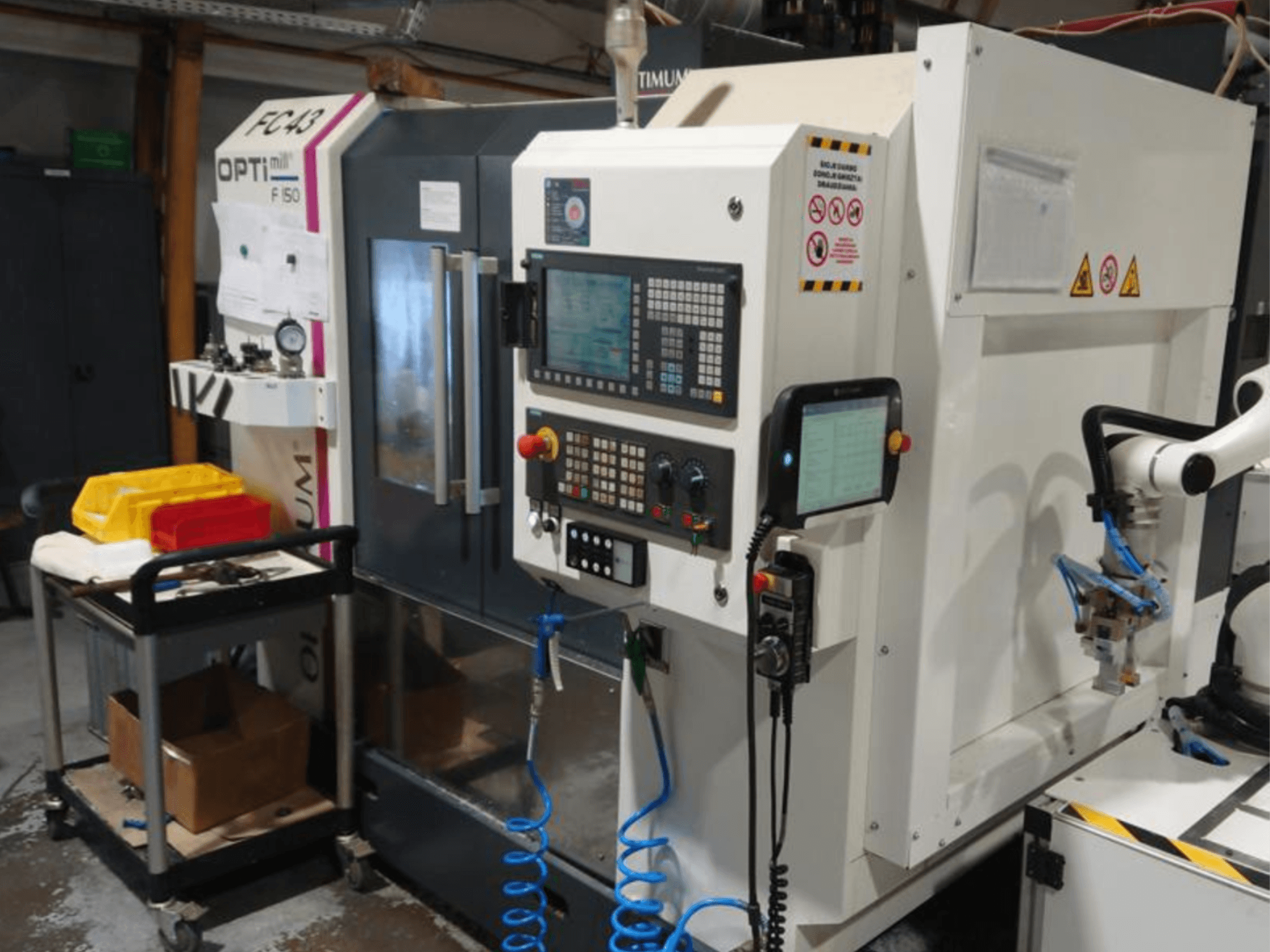Control panel of OPTIMUM F50 milling machine with hoses, tools, and a robotic arm visible in a workshop setting.