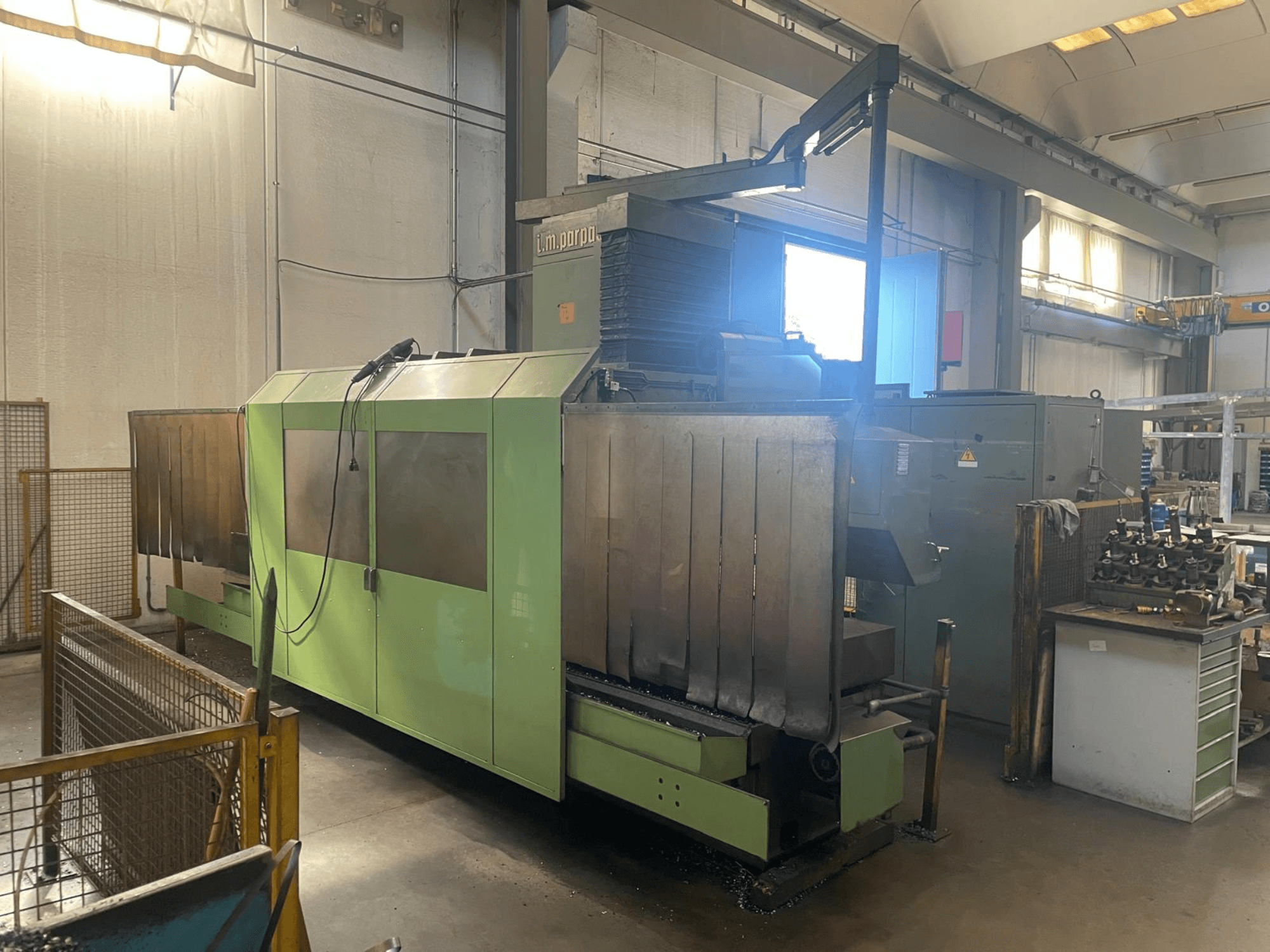 Green industrial machine with visible metal components, front view; features control panel, safety warnings, and workstation nearby.