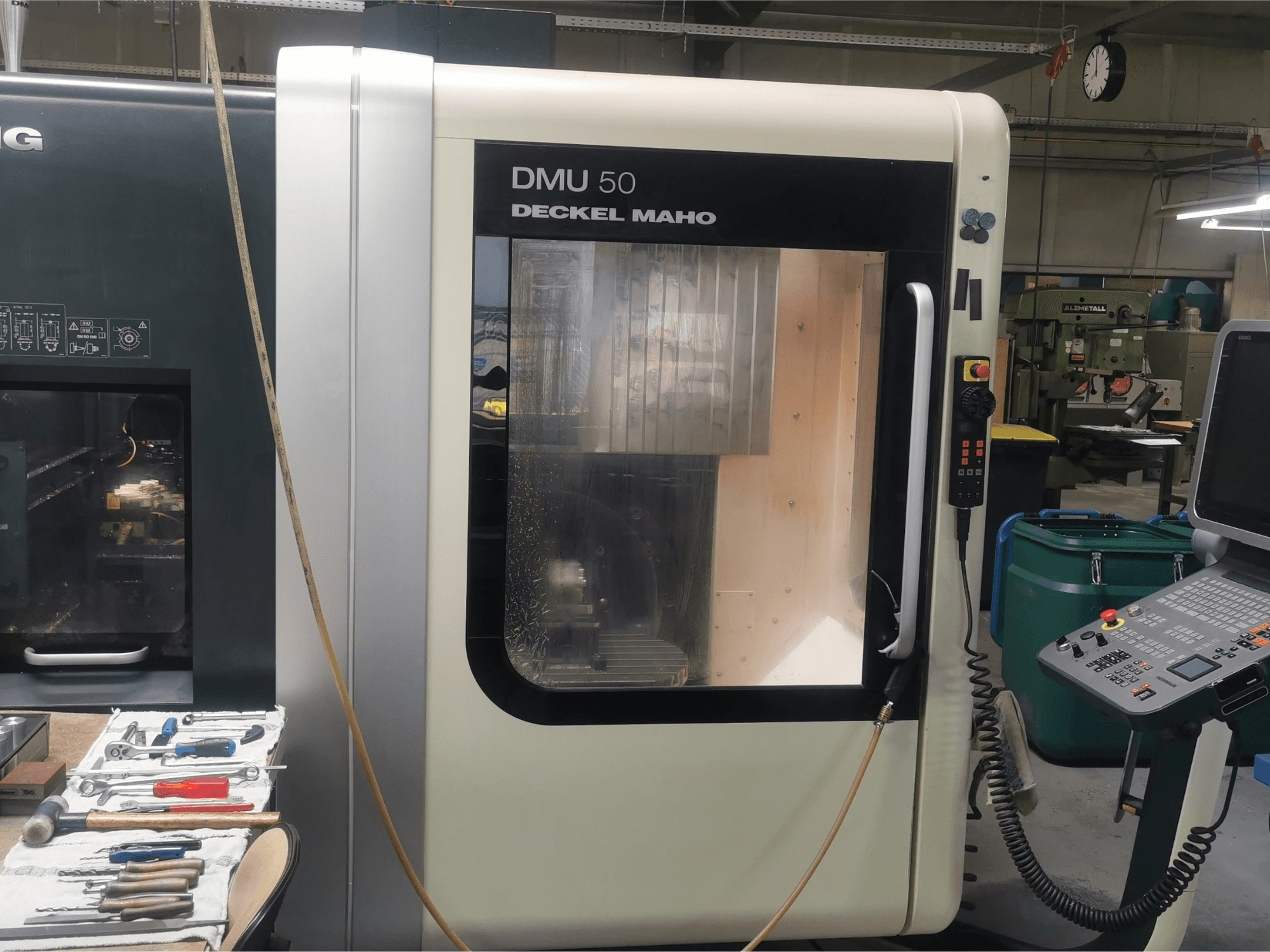 DMU 50 deckel maho CNC machine viewed from the front, showcasing its control panel and transparent door. Tools are nearby.