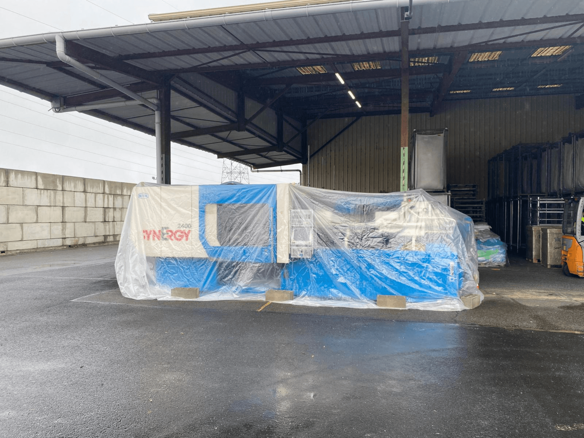Wrapped SYNERGY 2400 machine viewed from the front, blue and beige colors, inside a large covered industrial area.