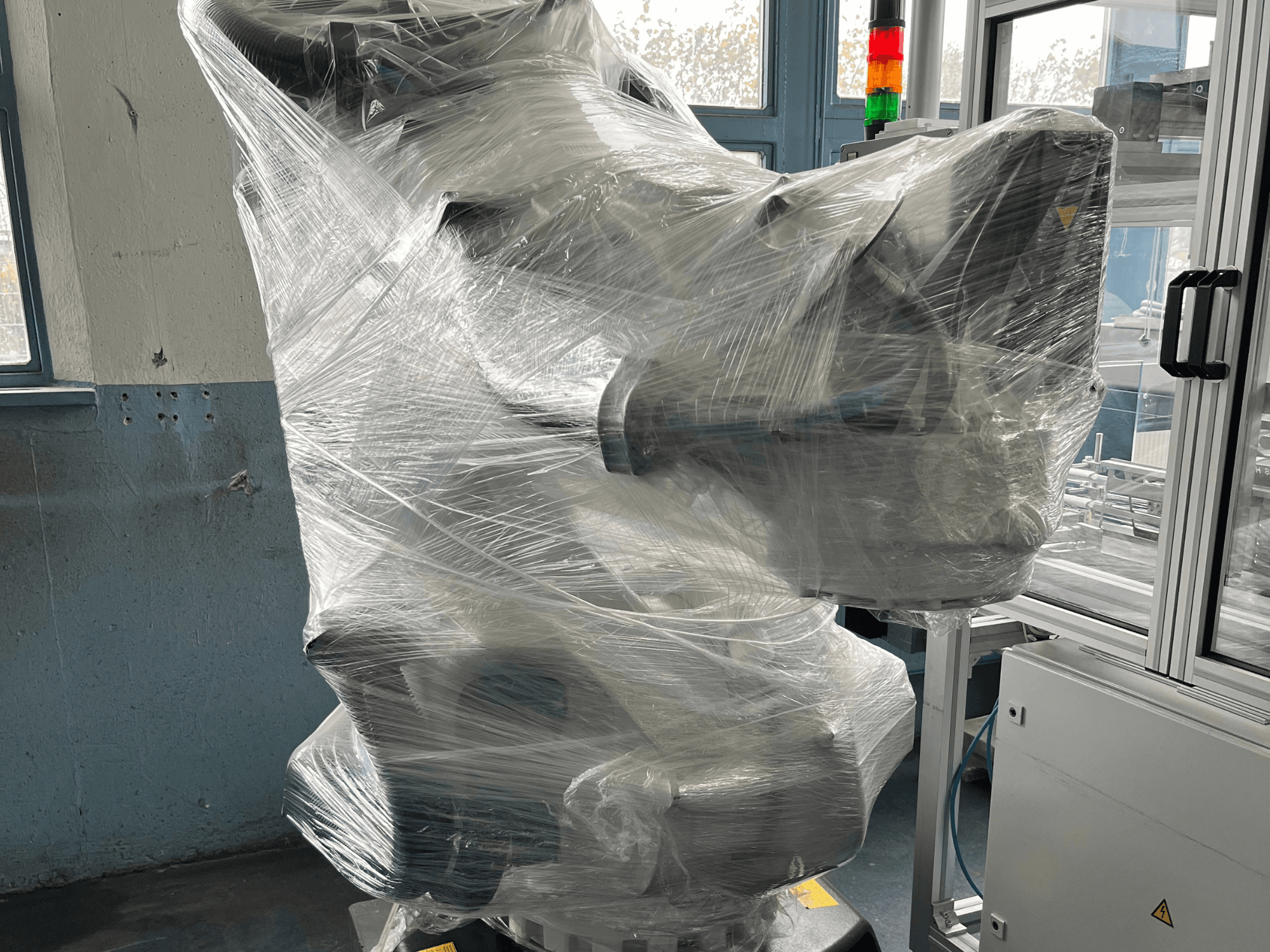 Industrial robotic arm wrapped in plastic, front view. Includes control unit and indicator light. Brand not visible.